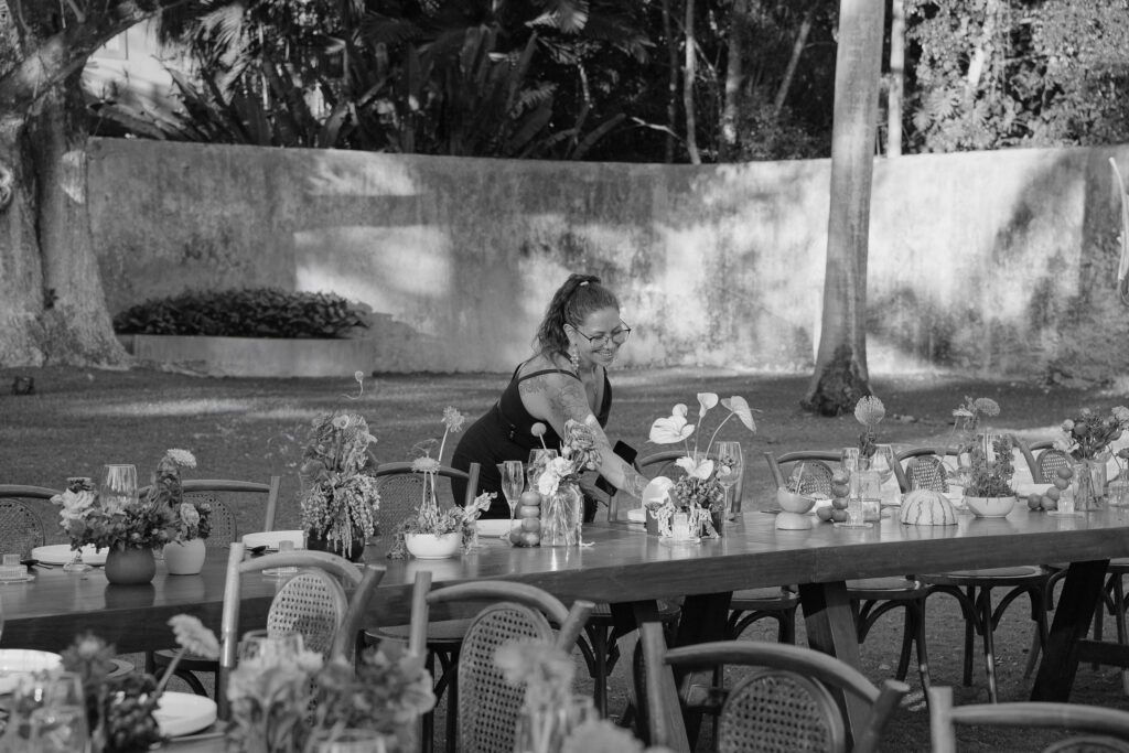 Joe arranging flowers and glassware along a long table before guests arrive