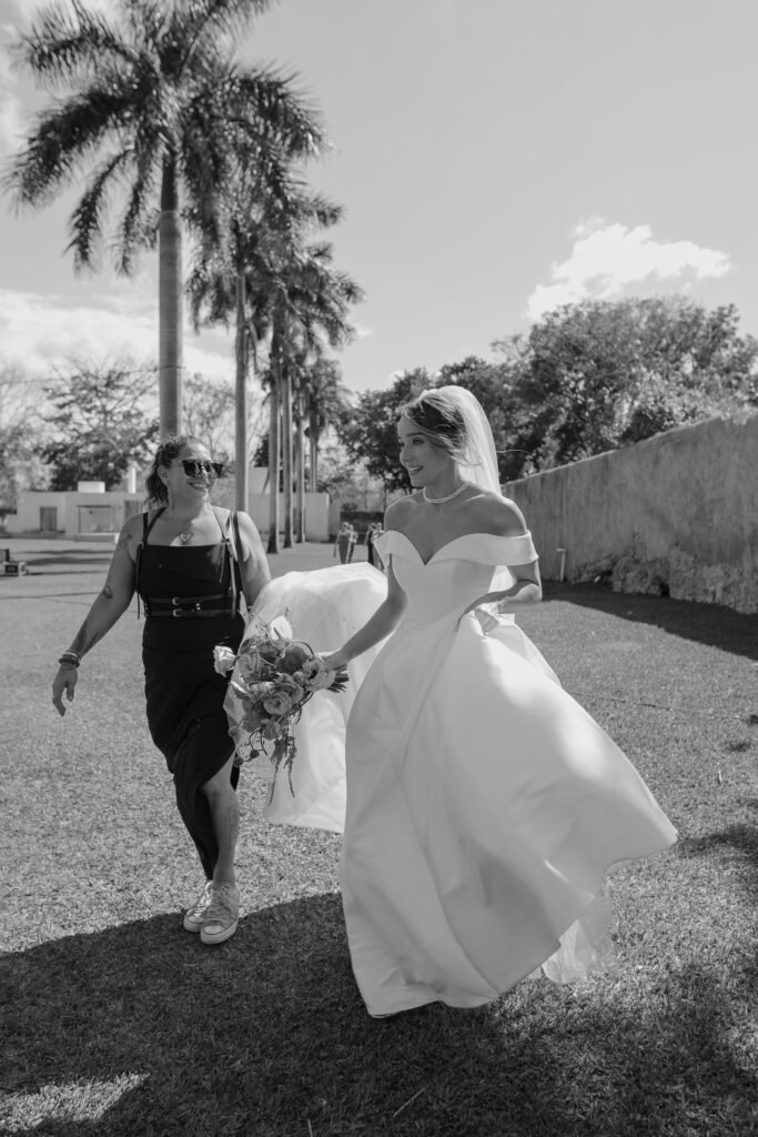 Joe walking beside the bride, carrying the train of her dress and laughing