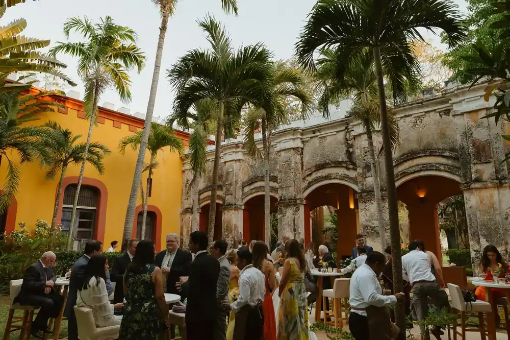 Guests at cocktail hour among palms and stone arches beside a yellow hacienda facade